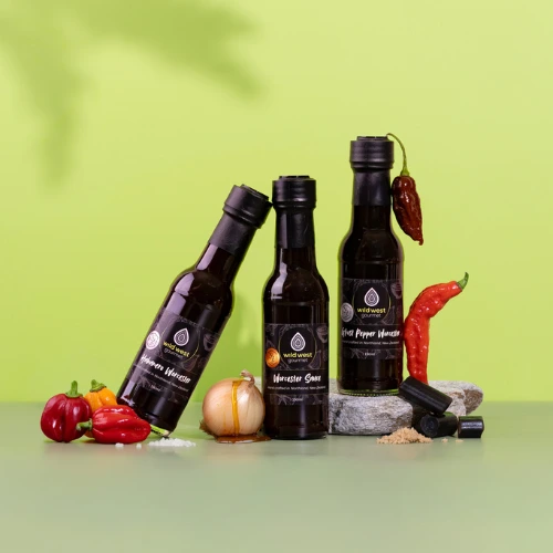 A line up of three bottles of Worcester sauce, with the left hand bottle leaning jauntily on the bottle to its right. There is a bright green backdrop.